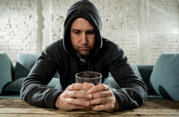 Мужчина в капюшоне сидит за столом, держит в руках стакан со спиртным