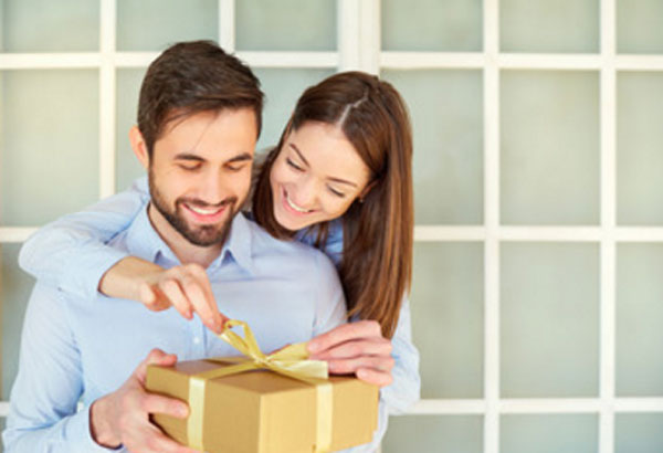 Женщина стоит за спиной мужчины и помогает ему открыть коробку с подарком