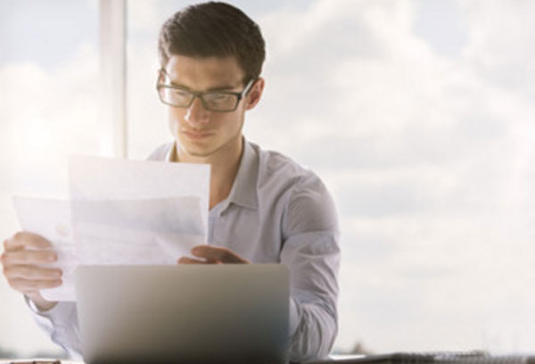Мужчина в очках сидит перед ноутбуком, рассматривает листки бумаги