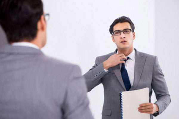 Мужчина в очках с докладом в руке стоит перед зеркалом и поправляет свой галстук от волнения