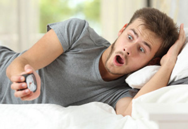Мужчина лежит в кровати с ужасом смотрит на будильник
