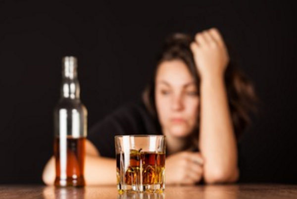 Замученная женщина, перед ней стоит бутылка и стакан со спиртным