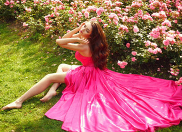 Девушка в вечернем ярко розовом платье сидит на земле возле клумбы с цветами