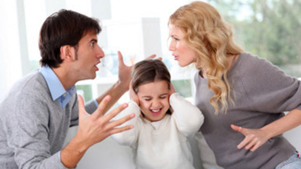 Девочка сидит между родителями, которые кричат друг на друга. Она закрывает руками уши.