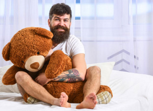 Инфантильный мужчина сидит на кровати, обнимая большую мягкую игрушку